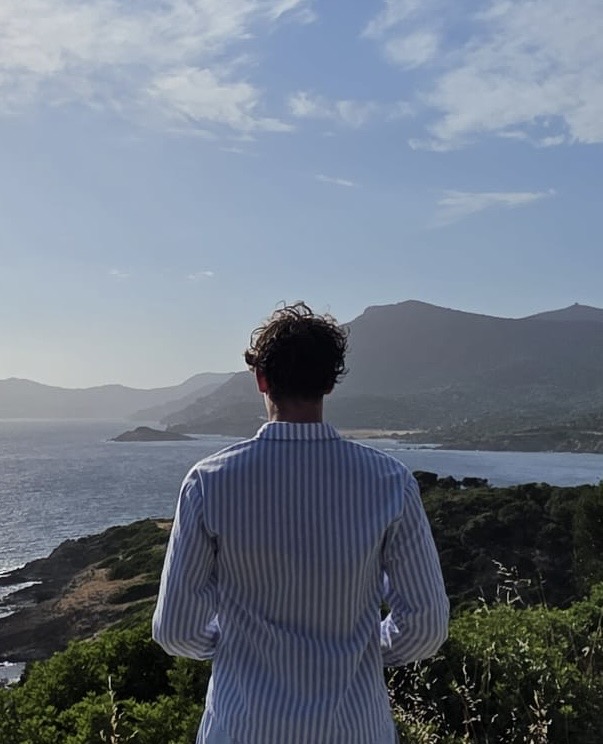 Gianluca de Bache von hinten, blickt über eine Küstenlandschaft mit Meer und Bergen im Abendlicht.
